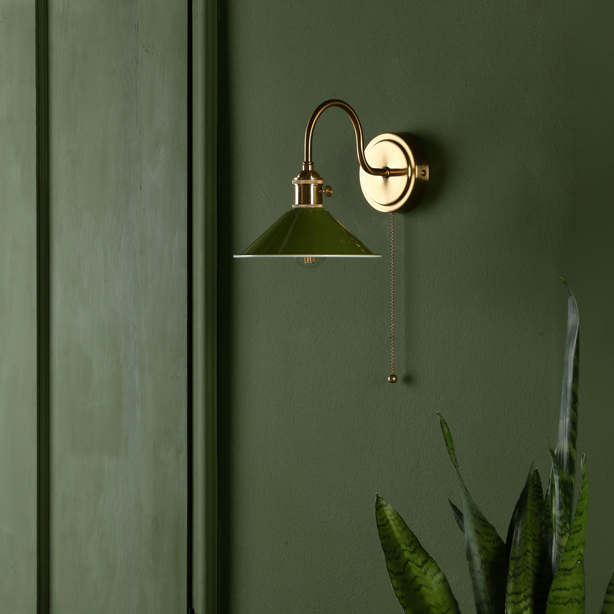 A brass wall sconce with a green metal shade and pull chain, mounted on a dark green wall.