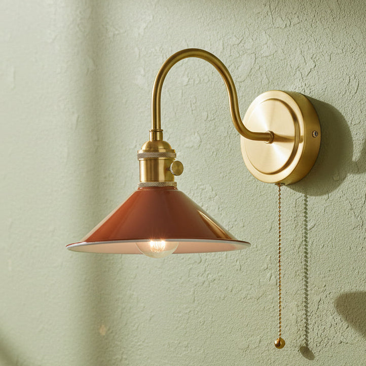 A brass wall sconce with a curved arm and a rust-colored, conical shade, featuring a pull chain.