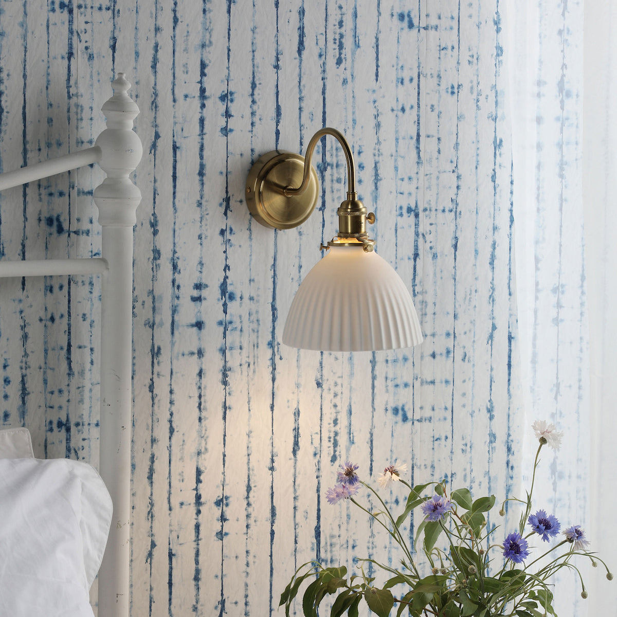 A brass wall sconce with a white fluted shade illuminates a bedroom with blue striped wallpaper.