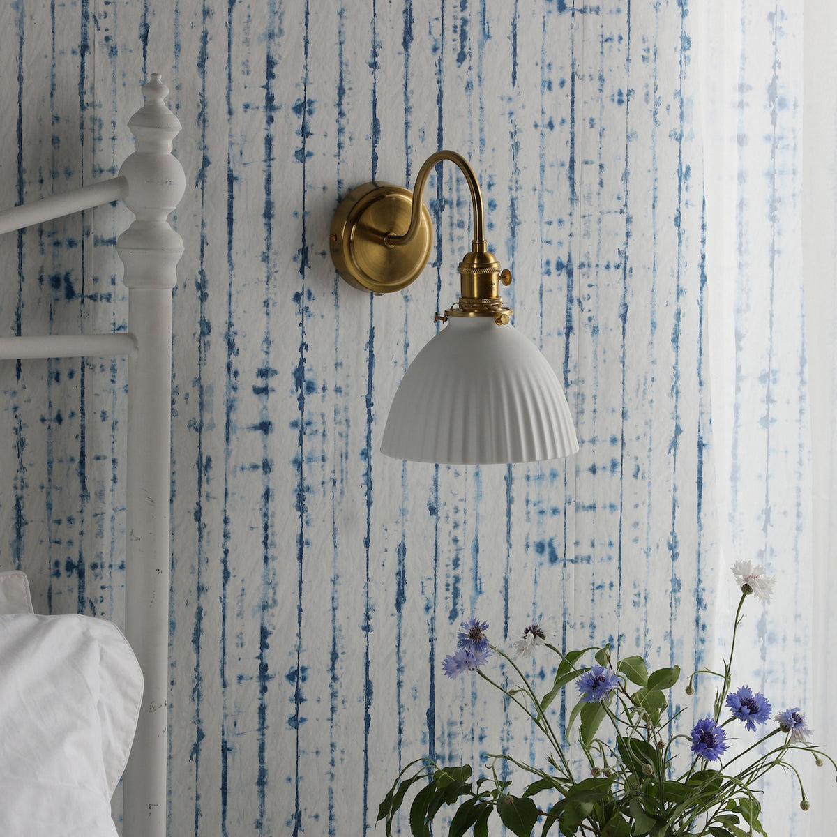 A white bed frame and a brass wall sconce with a white shade against blue and white striped wallpaper.