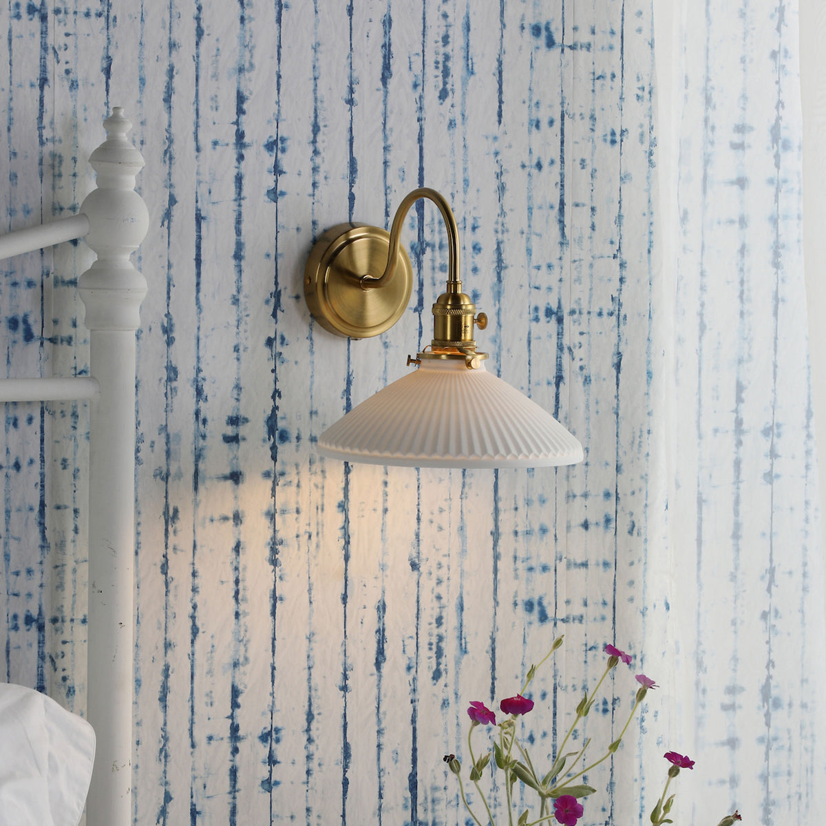 A brass wall sconce with a white fluted shade illuminates a blue and white striped wallpaper. Pink flowers are in the foreground.