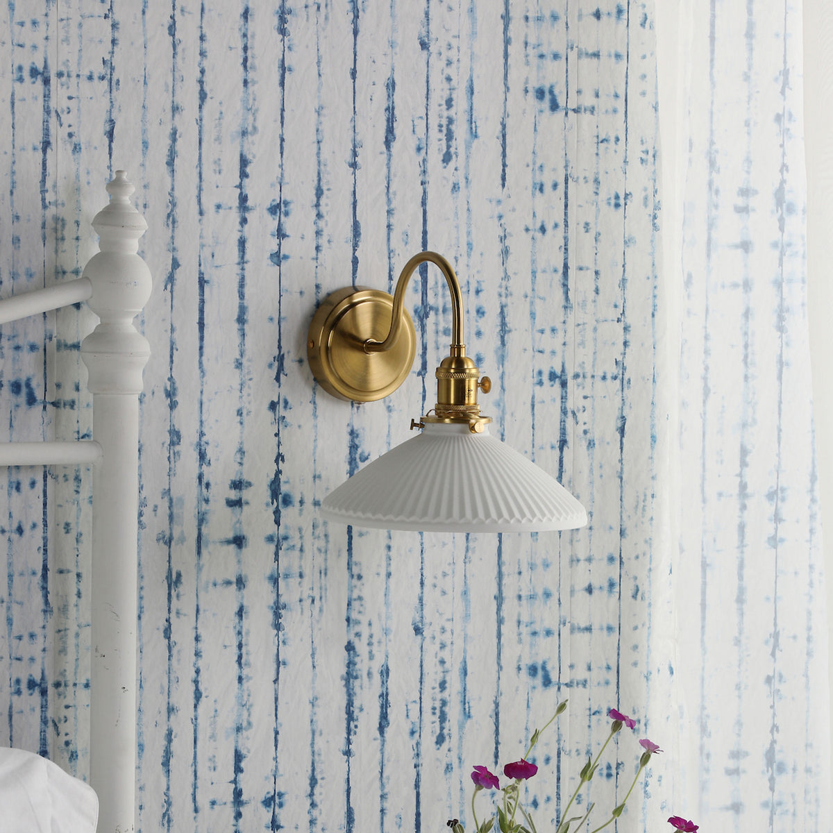 A brass wall sconce with a white fluted shade against blue and white striped wallpaper.