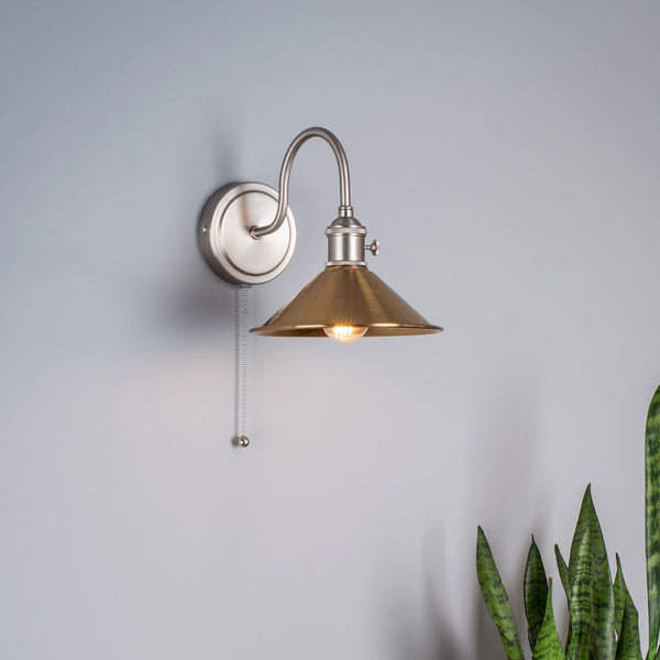 Brushed steel wall light with a brass conical shade and a pull chain switch. A green plant is visible on the right.