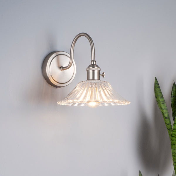 Brushed nickel wall sconce with a clear, fluted glass shade and an exposed bulb, next to a snake plant.