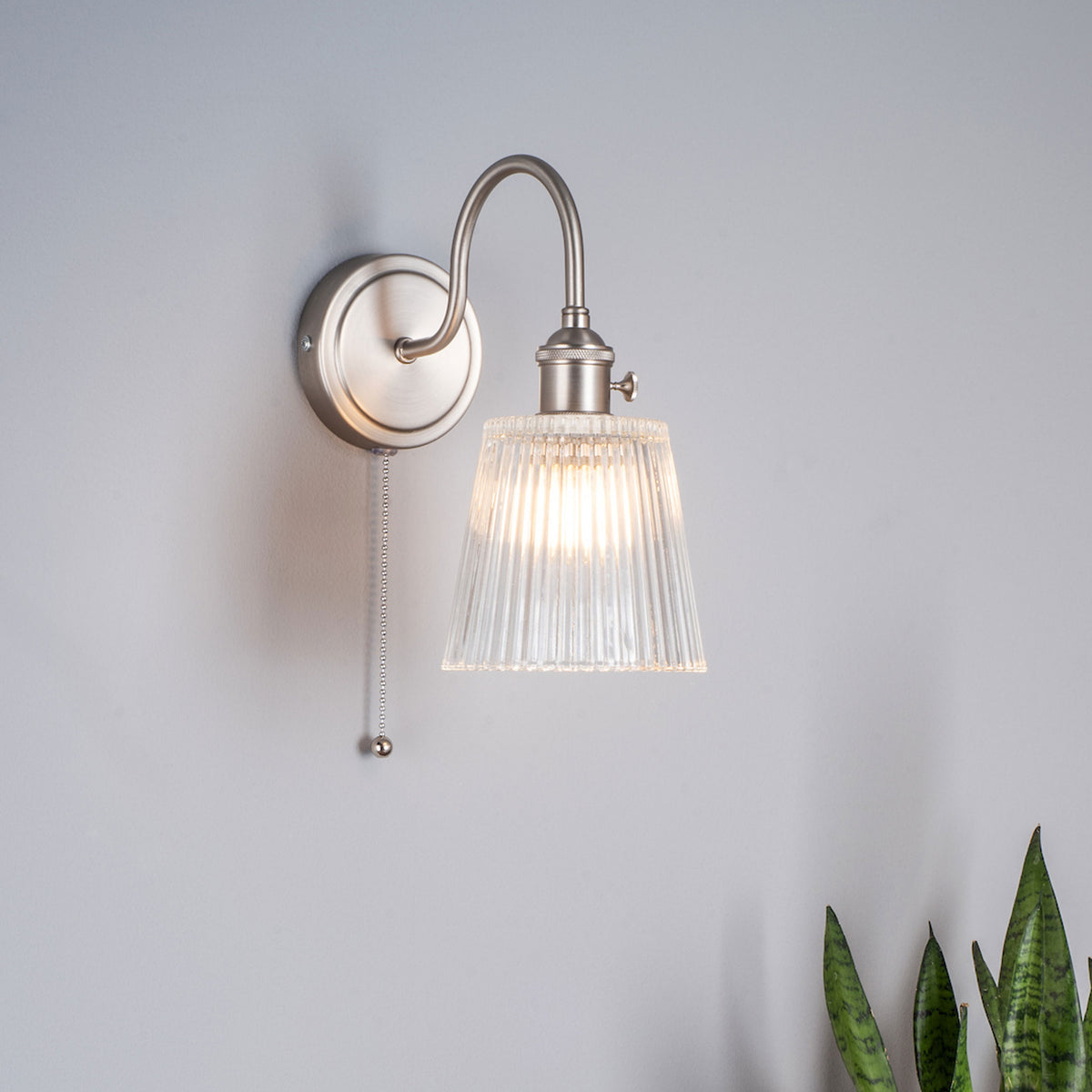 Brushed chrome wall light with a fluted glass shade and a pull cord switch. It emits a warm glow.