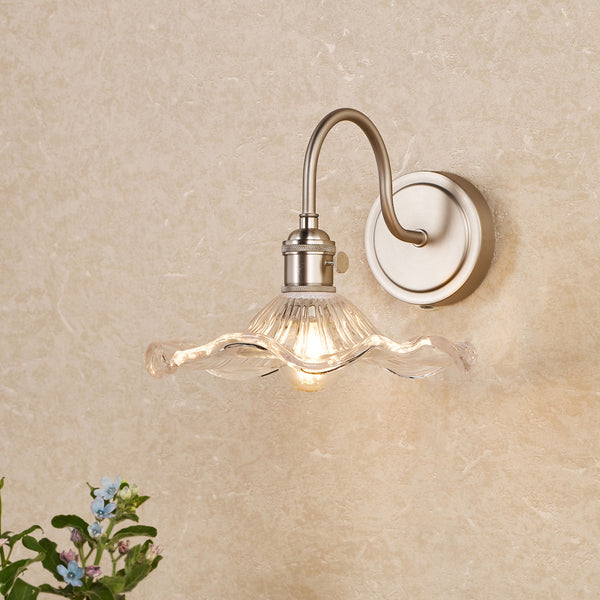 A wall-mounted light fixture with a brushed nickel finish and a clear, ruffled glass shade. It emits a warm glow.