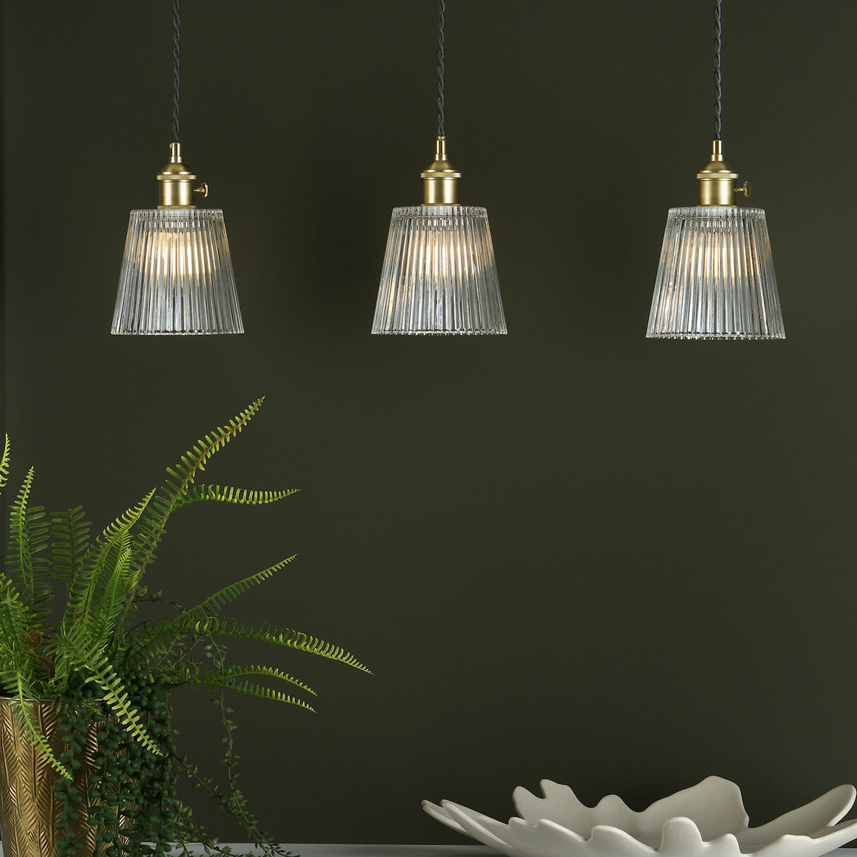 Three pendant lights with ribbed glass shades and brass fittings hang against a dark green wall. A fern is in a gold pot.