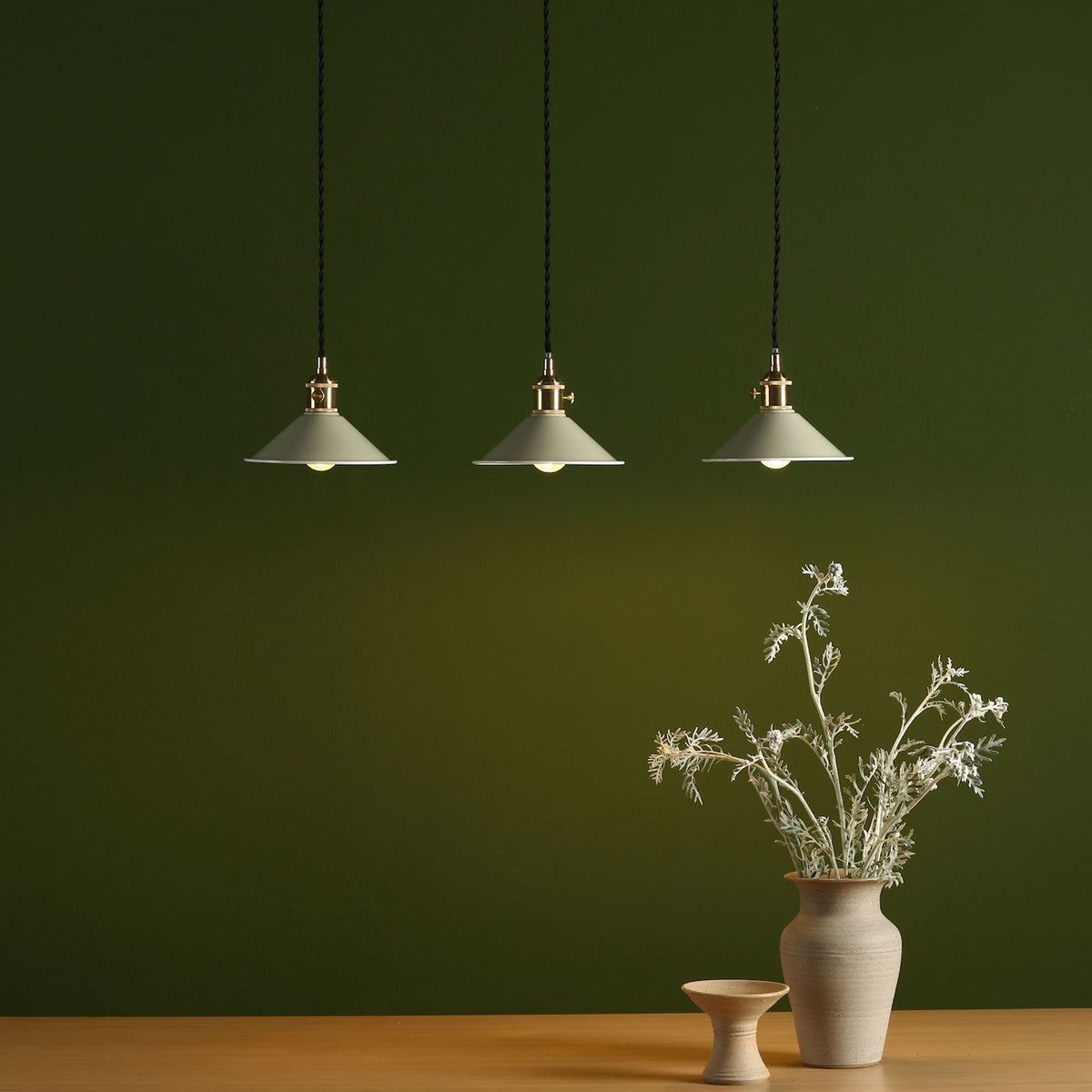Three pendant lights with light green shades hang from a dark green ceiling. A vase of dried flowers sits on a table.