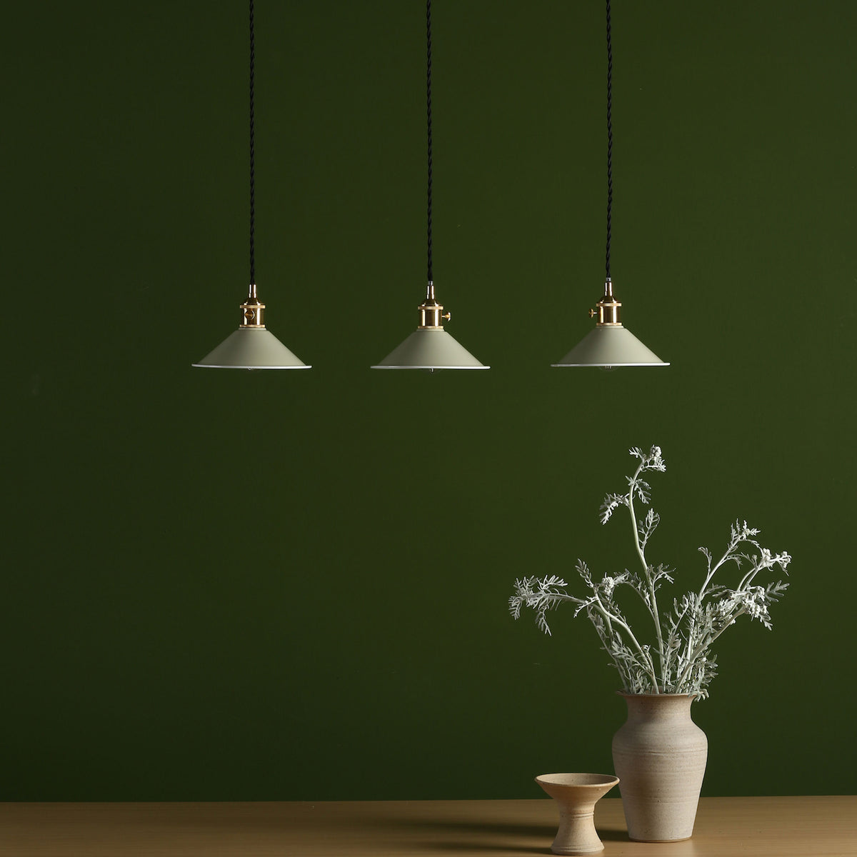 Three pendant lights with pale green shades hang above a wooden table with a vase of white foliage.