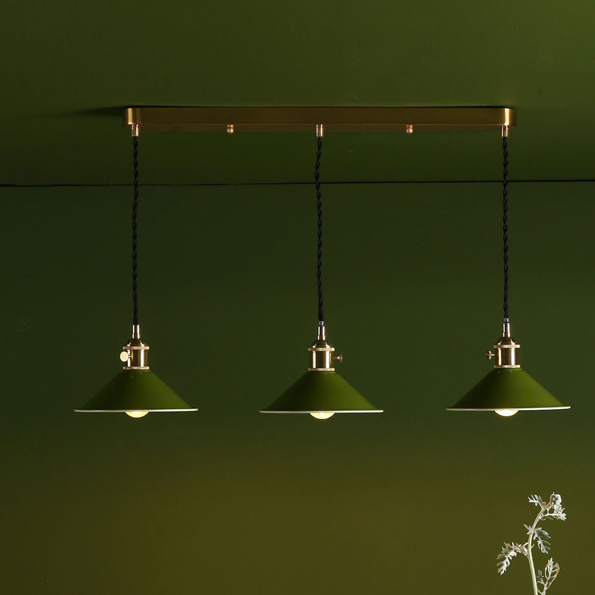 Three green pendant lights with brass fittings hang from a gold bar against a dark green wall.