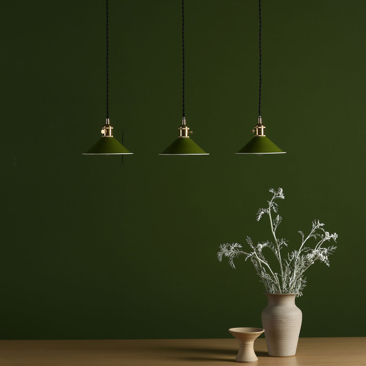 Three green pendant lights with brass fittings hang against a dark green wall. A vase of dried flowers sits on a table.