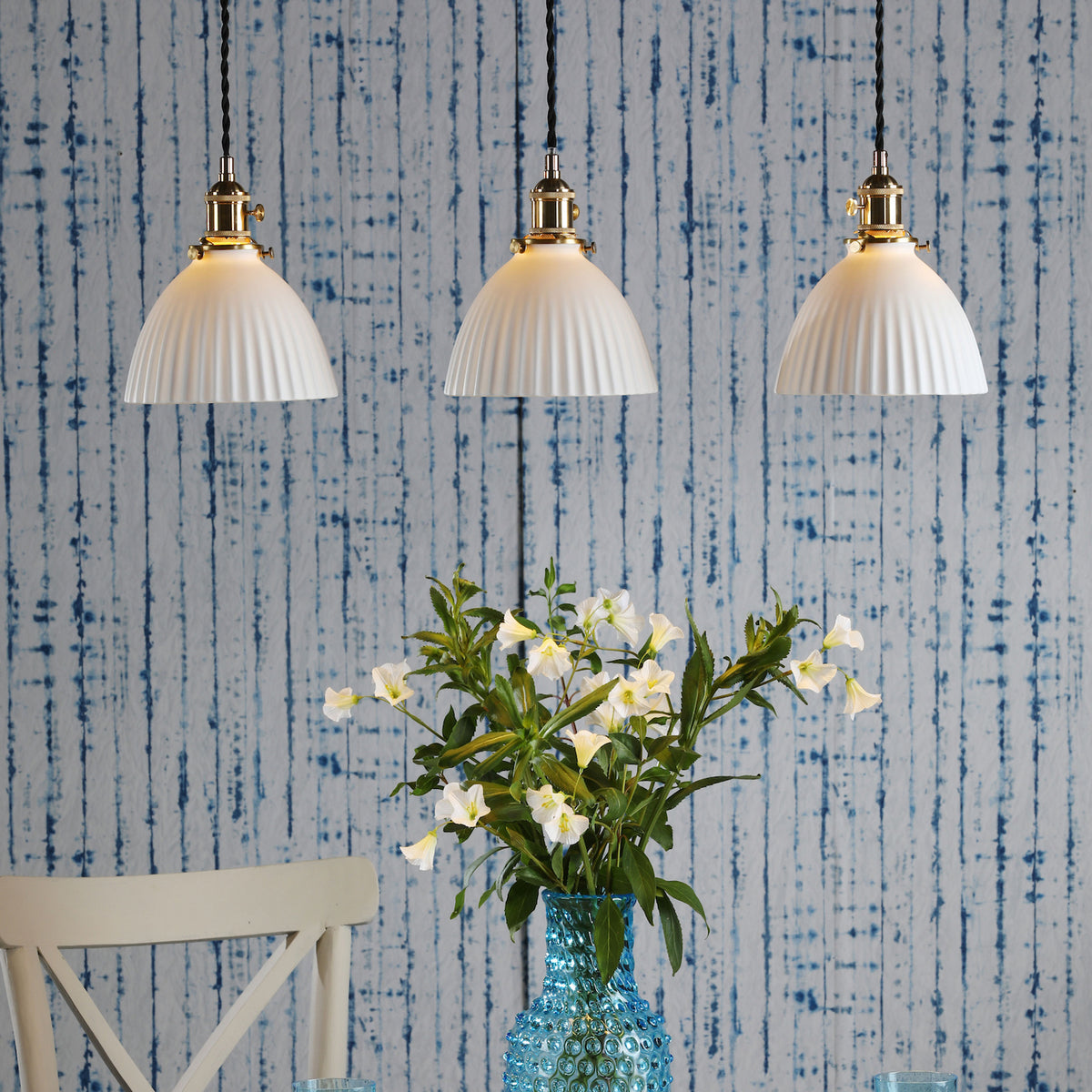 Three white fluted pendant lights hang above a blue hobnail vase with white flowers. Blue striped wallpaper.