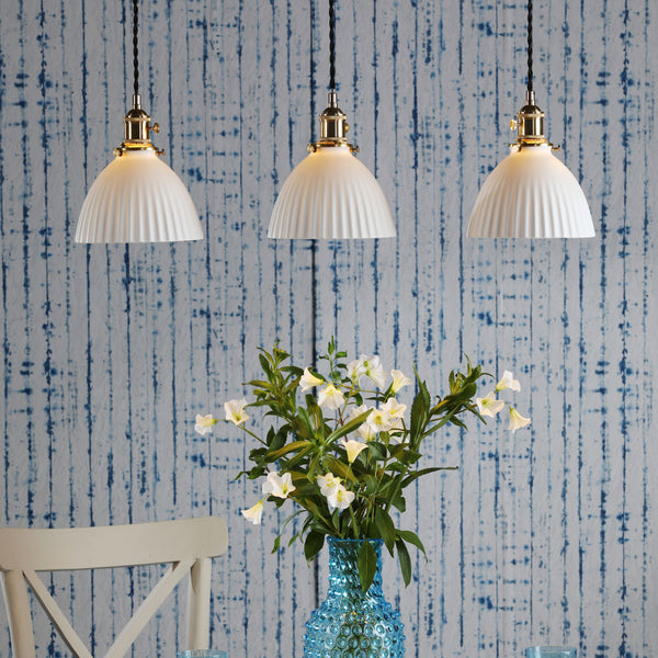 Three white fluted pendant lights hang above a blue hobnail vase with white flowers. Blue striped wallpaper.
