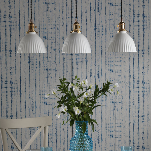 Three white fluted pendant lights hang above a blue textured vase with white flowers. Blue striped wallpaper.