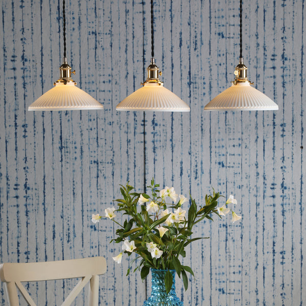 Three pendant lights with pleated white shades hang above a vase of white flowers and a chair.