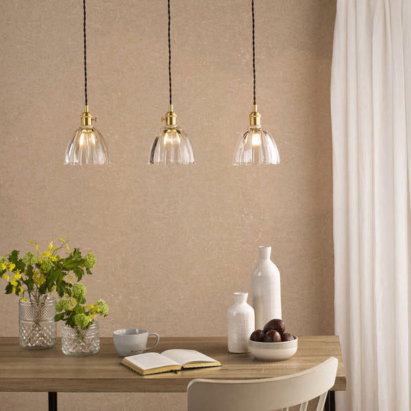 Three clear glass pendant lights with brass fittings hang above a wooden table with flowers, books, and decorative vases.