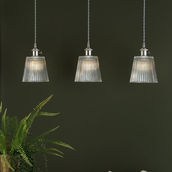 Three ribbed glass pendant lights with brushed nickel fittings hang against a dark green wall, with a fern in the foreground.