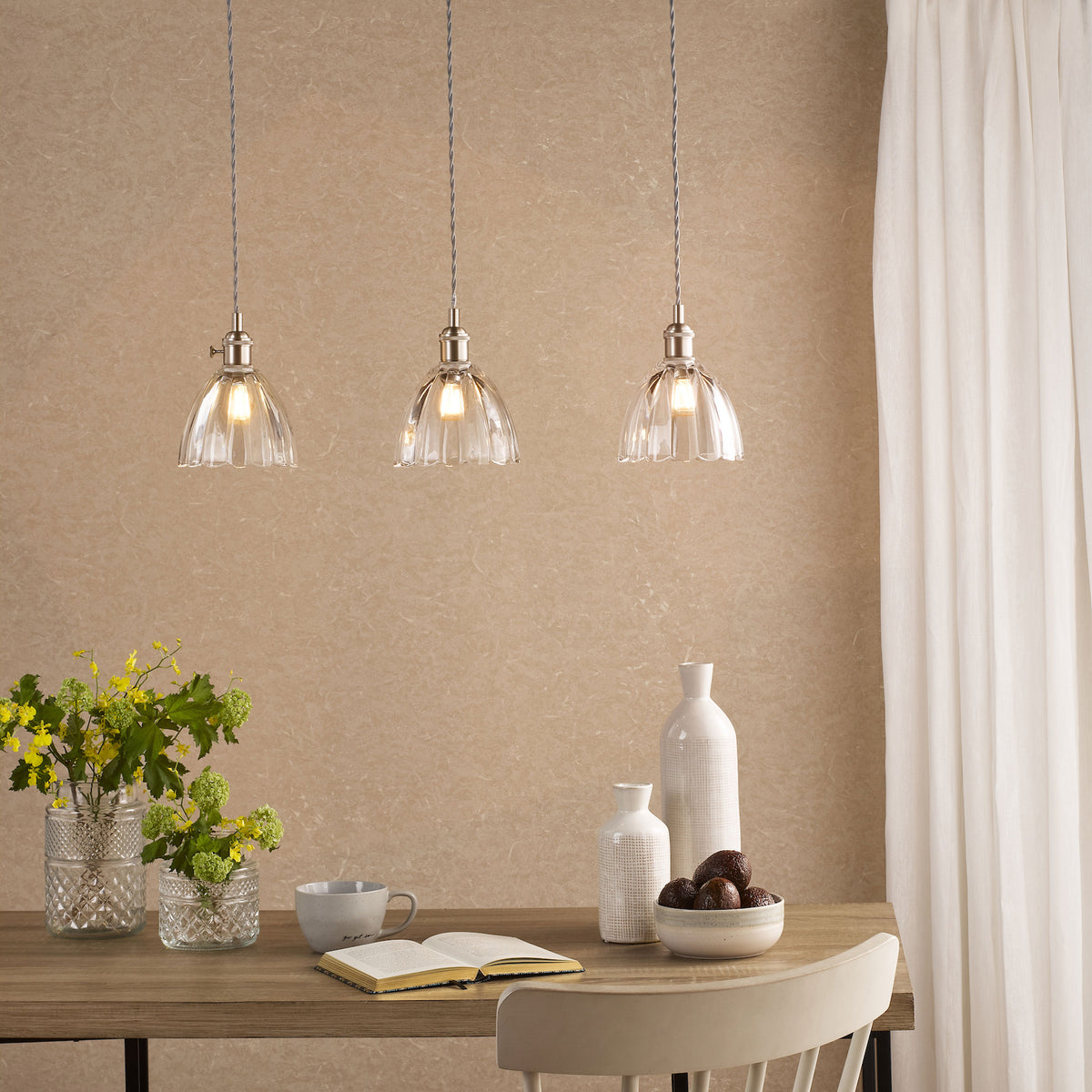 Three clear glass pendant lights with brushed nickel hardware illuminate a wooden table with flowers, a book, and fruit.