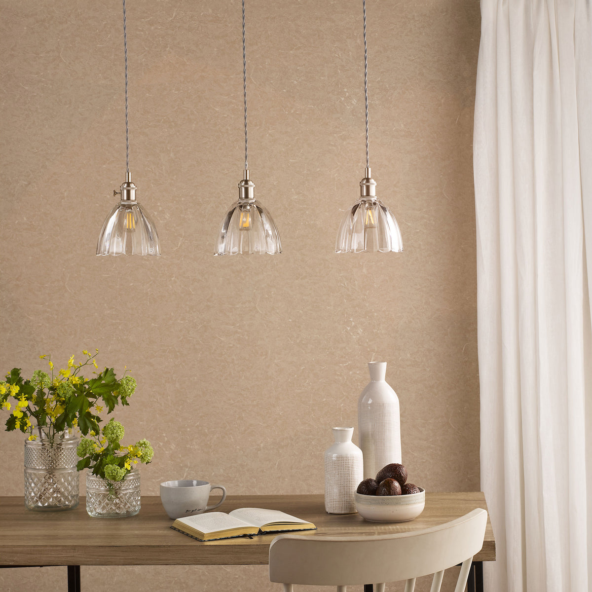 Three clear glass pendant lights hang above a wooden table with flowers, books, and decorative items.