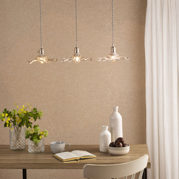 Three glass pendant lights hang above a wooden table with flowers, vases, a book, and a bowl of fruit.