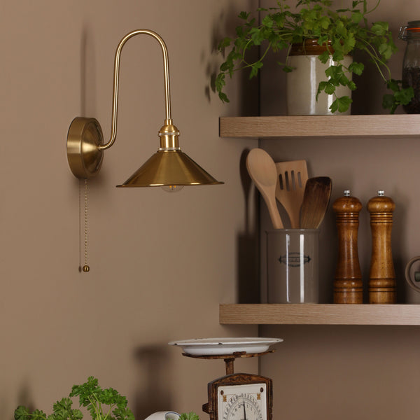 A brass wall light with a conical shade and pull chain, illuminating a kitchen corner with shelves and herbs.
