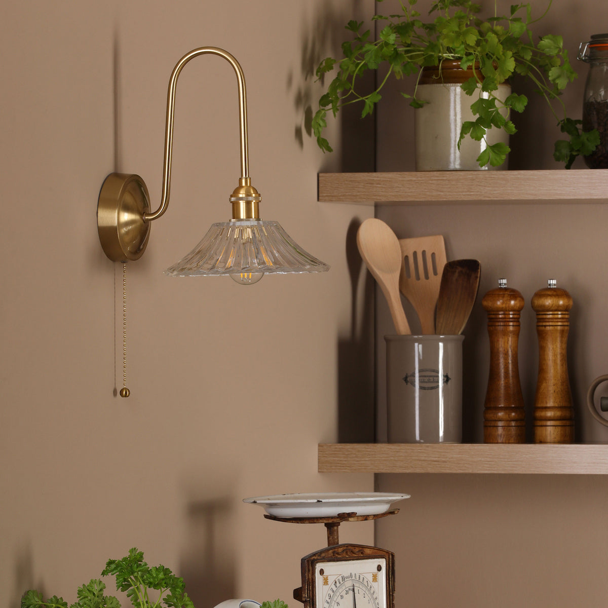 A brass wall light with a fluted glass shade illuminates a kitchen shelf with utensils and spices.