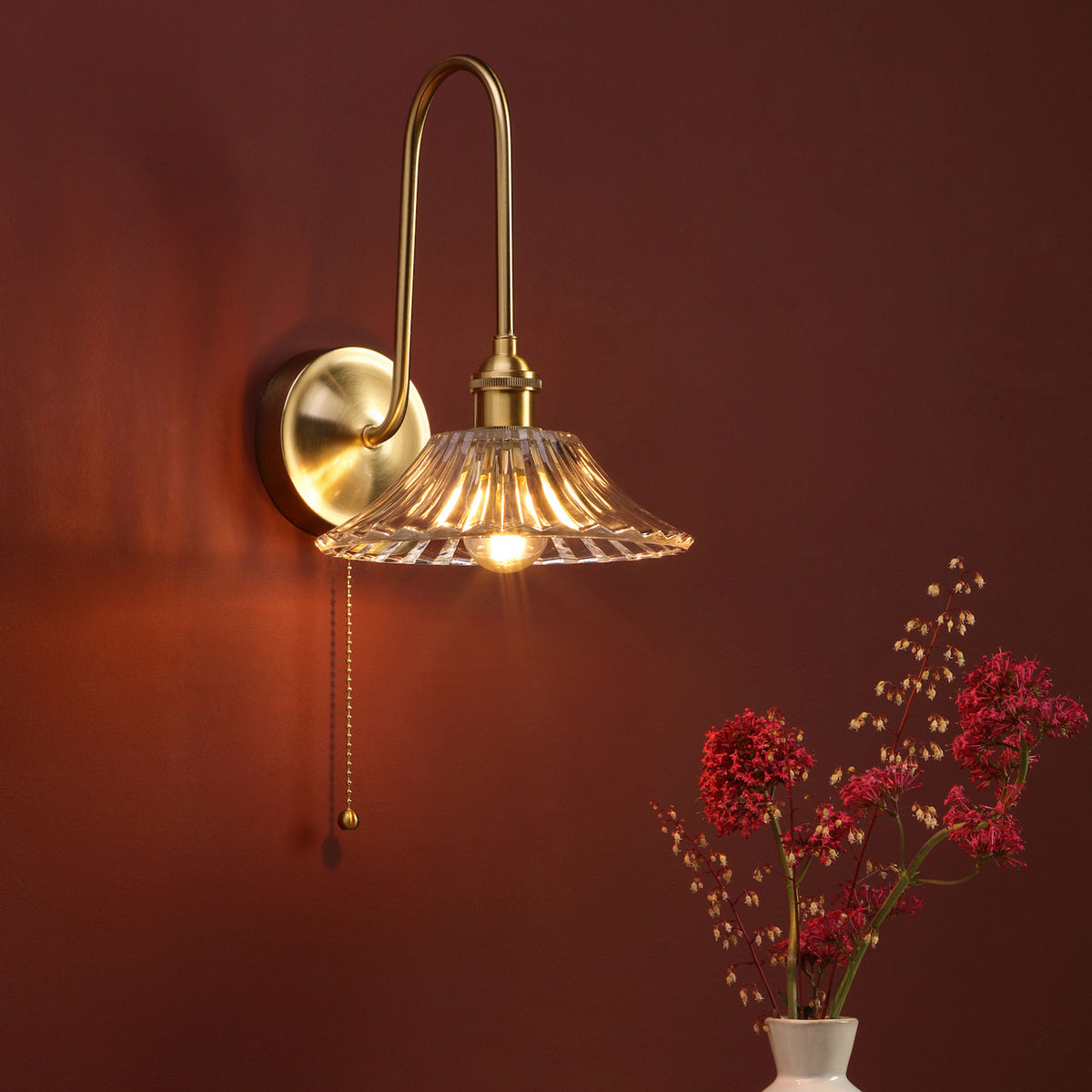 A brass wall sconce with a fluted glass shade illuminates a dark red wall, next to a vase of red flowers.