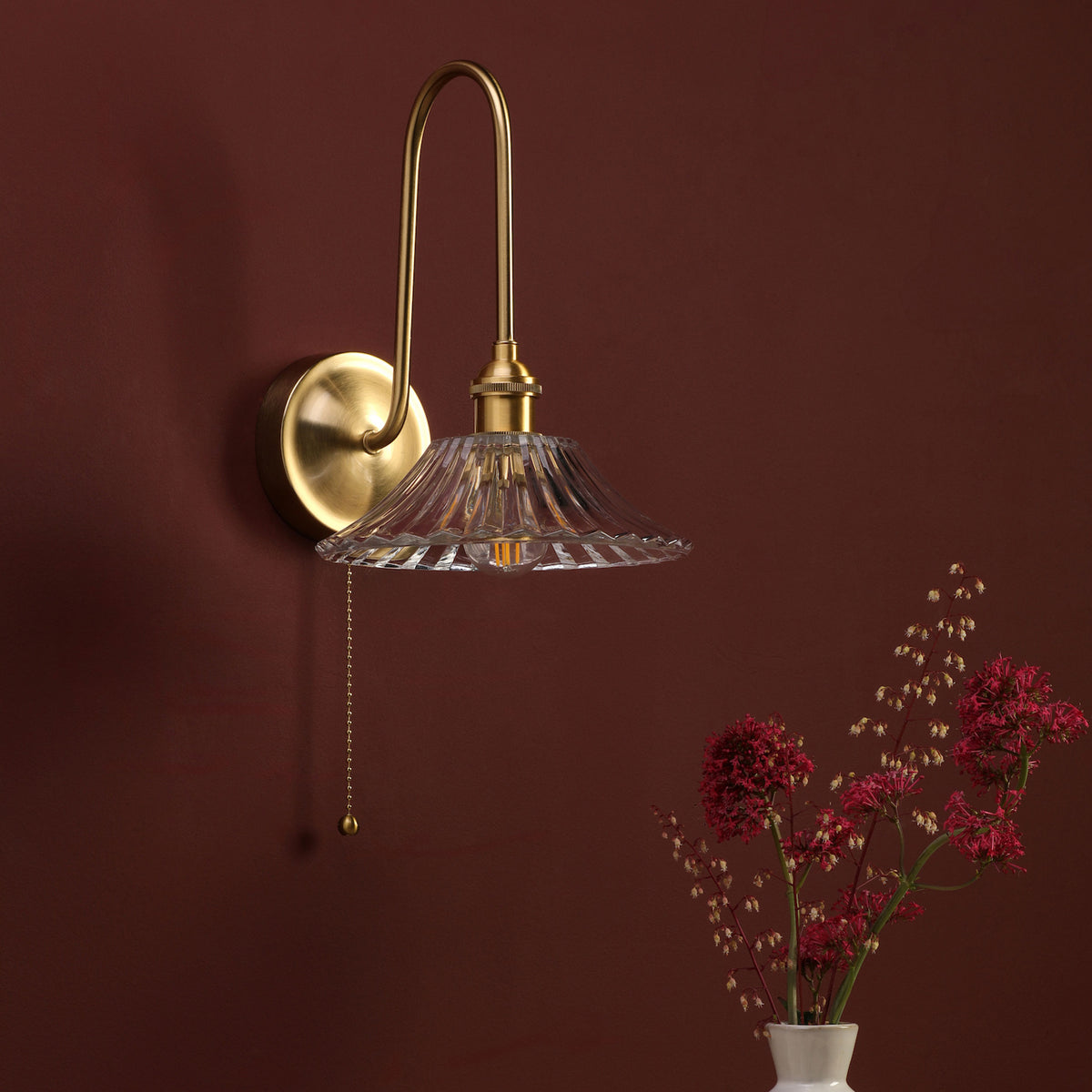 Brass wall sconce with a fluted glass shade and pull chain, mounted on a deep red wall with flowers.