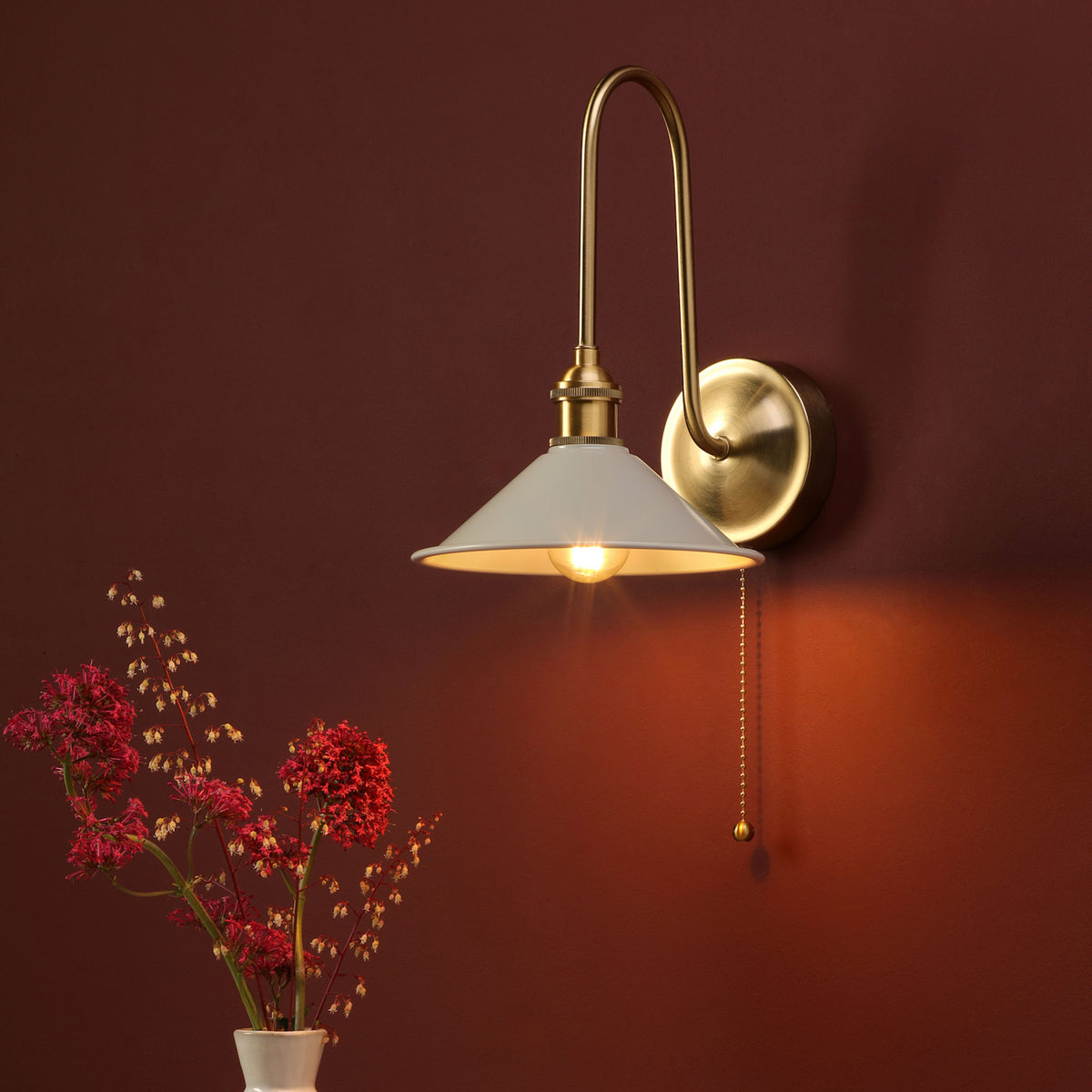 A brass wall sconce with a white shade and pull chain, casting a warm glow on a deep red wall, next to a vase of red flowers.