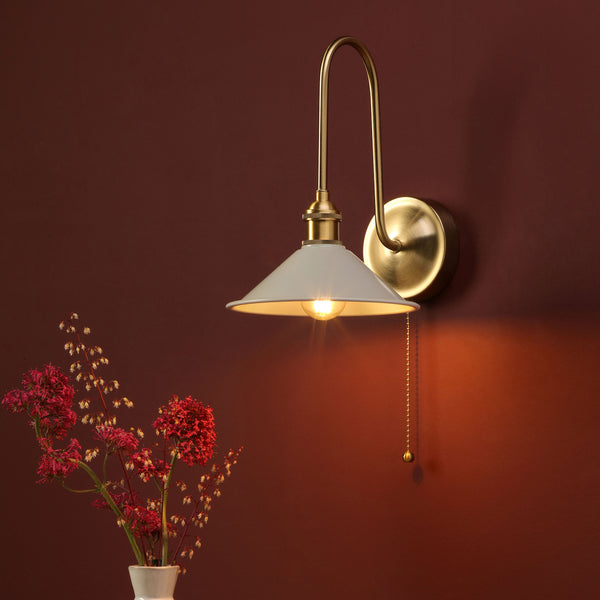 A brass wall sconce with a white shade and pull chain, casting a warm glow on a deep red wall, next to a vase of red flowers.