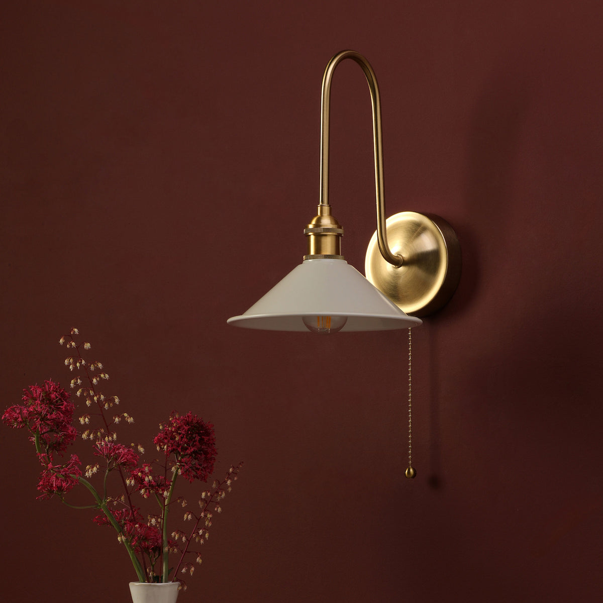 Brass wall light with a white shade and pull chain, next to a vase of red flowers.