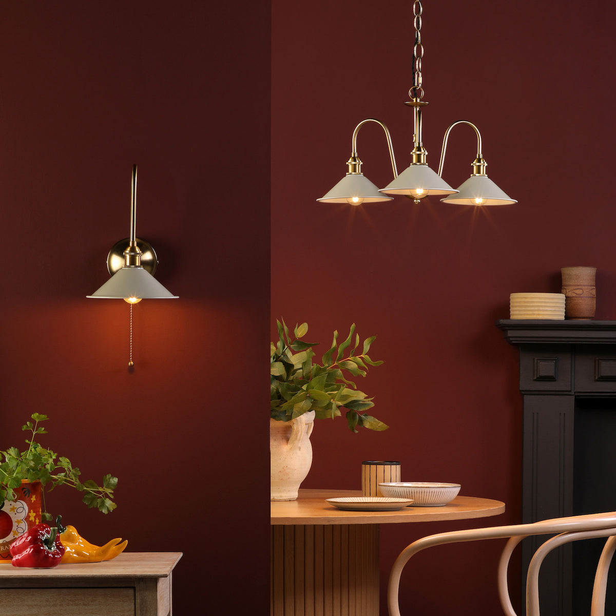 A brass wall light and a three-light chandelier with white shades illuminate a deep red room.