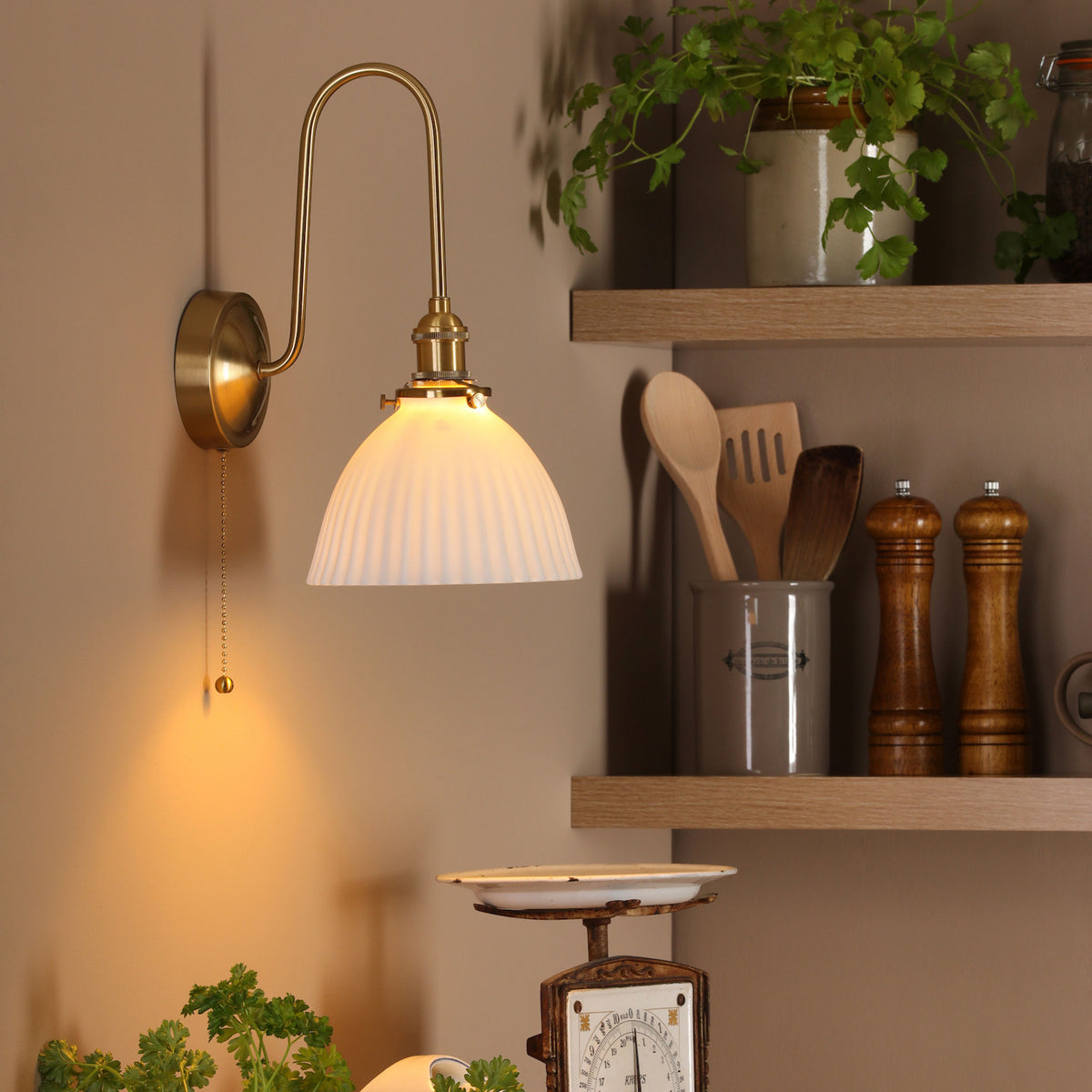A brass wall light with a ribbed glass shade illuminates a kitchen wall above an antique scale.