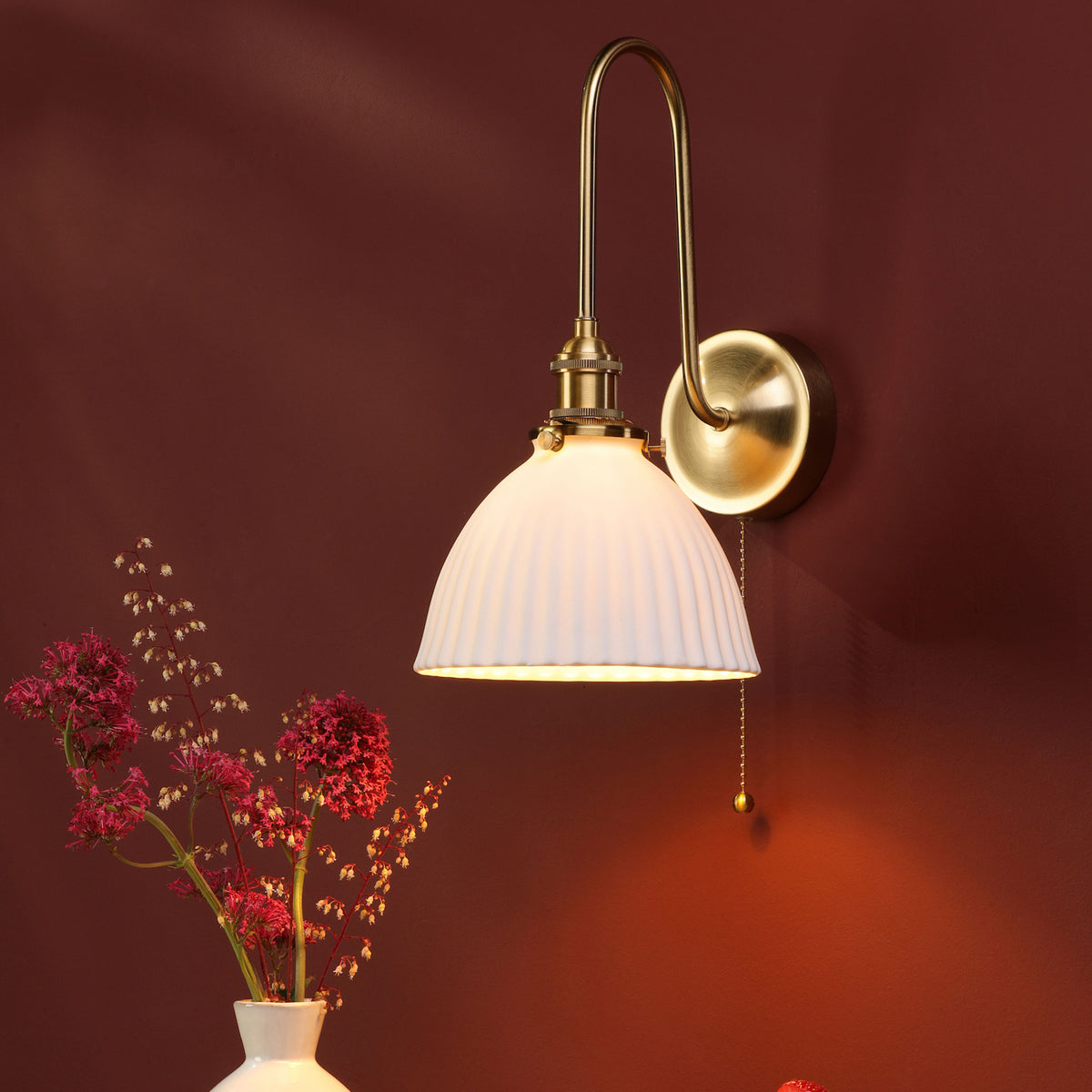 A brass wall sconce with a white fluted shade illuminates a dark red wall and a vase of flowers.