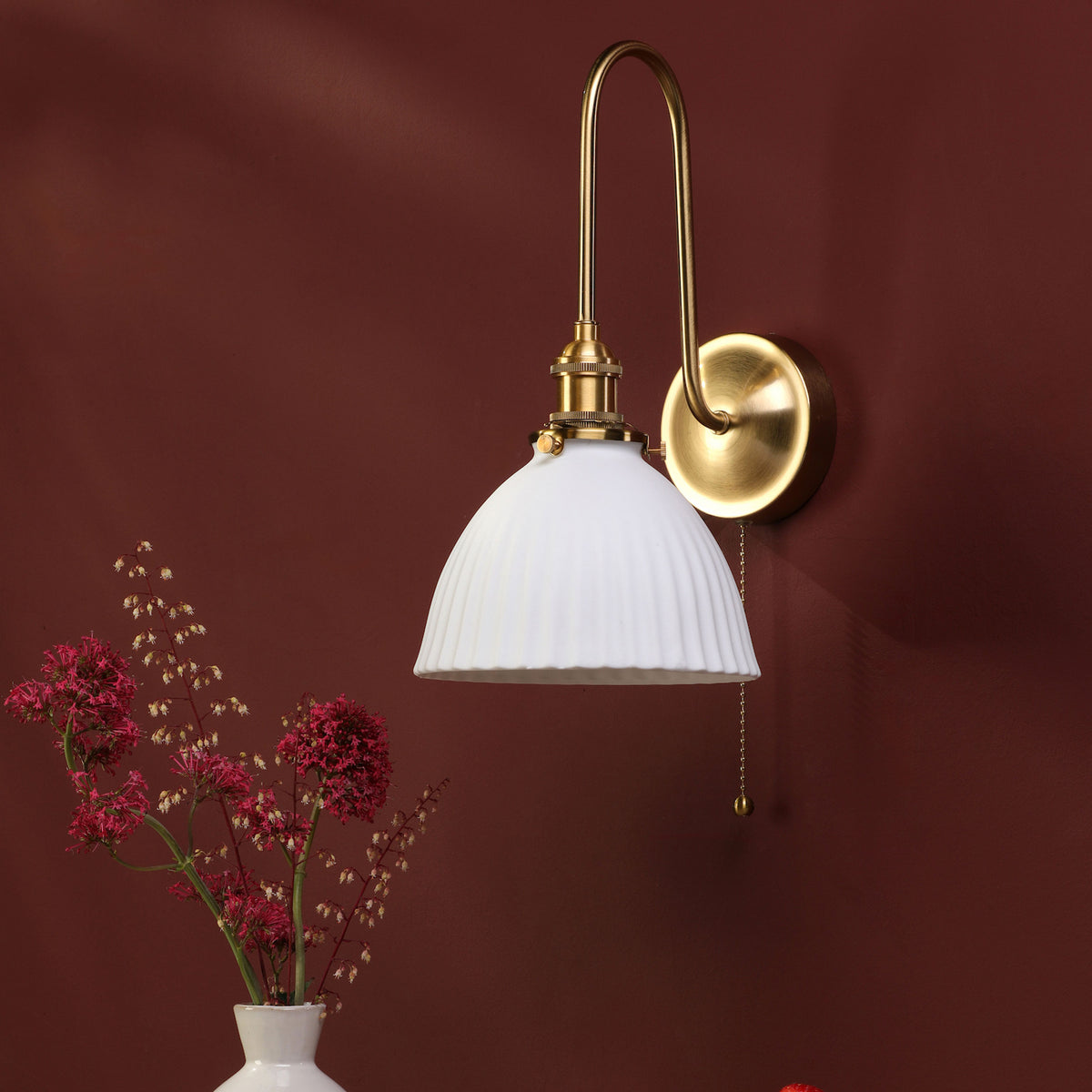 A brass wall sconce with a white fluted shade and pull chain, mounted on a deep red wall. Red flowers in a white vase.
