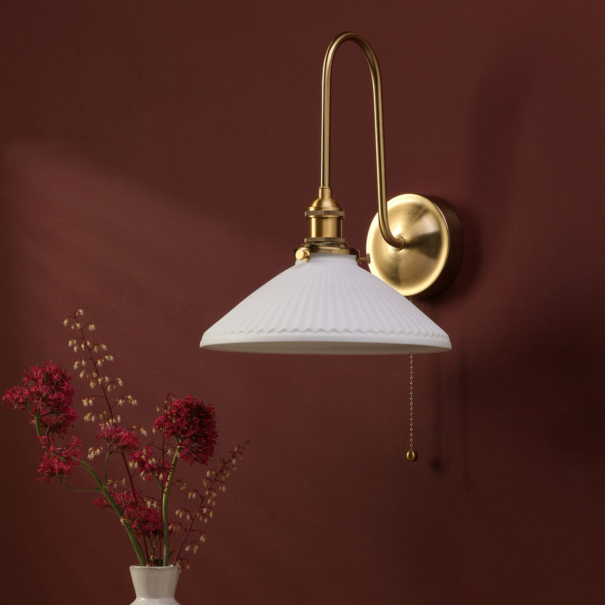 Brass wall sconce with a white fluted shade and pull chain, next to a vase of red flowers.