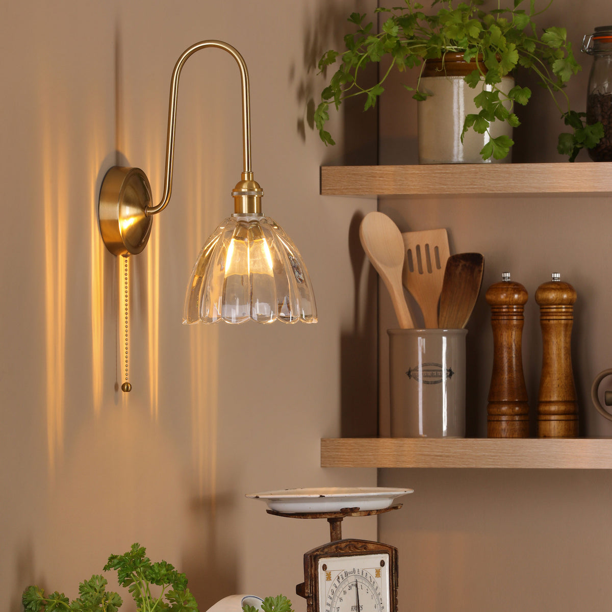 A brass wall light with a fluted glass shade illuminates a kitchen corner with wooden shelves and herbs.