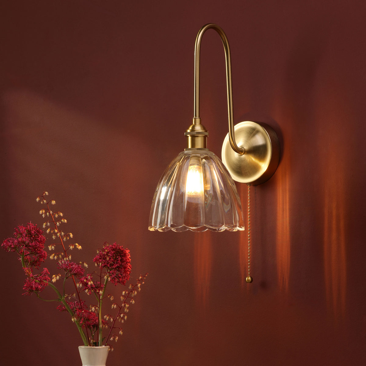A brass wall sconce with a fluted glass shade illuminates a dark red wall, next to a vase of red flowers.