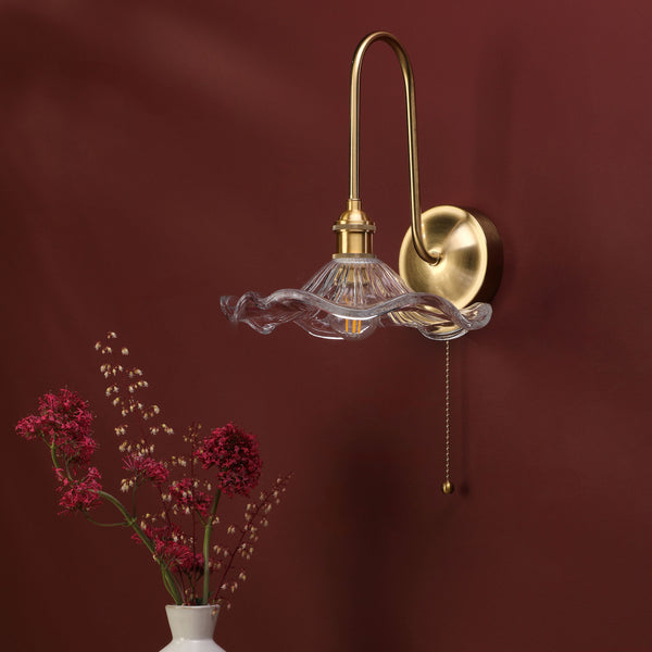 Brass wall sconce with a ruffled glass shade and a pull chain, next to a vase of red flowers.