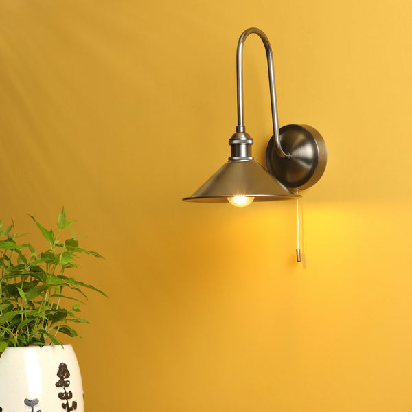 A brushed nickel wall sconce with a conical shade and a pull chain, illuminating a yellow wall above a plant.