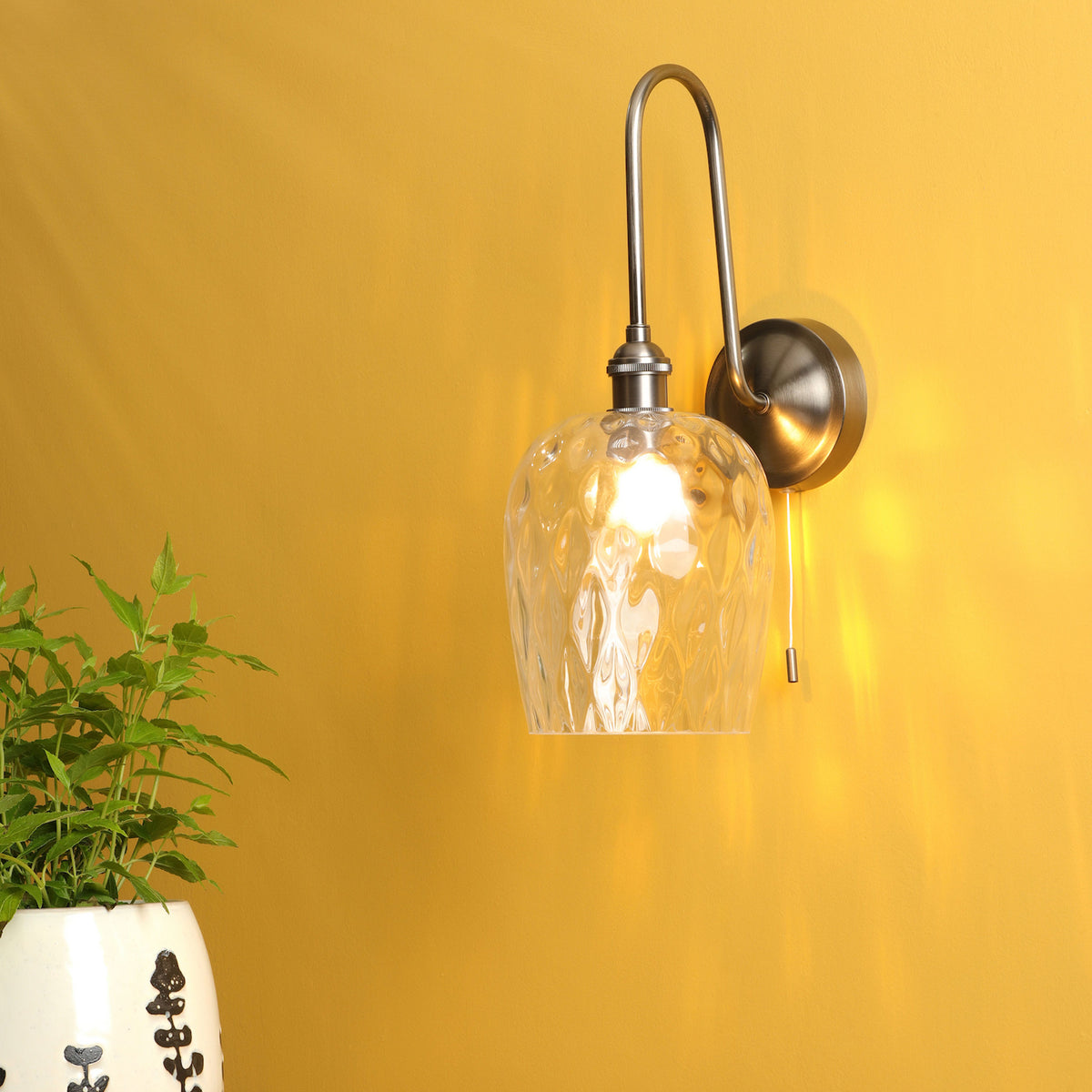 A wall sconce with a clear, textured glass shade and a brushed metal arm, illuminating a yellow wall.