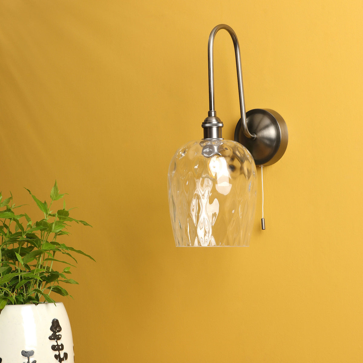 A wall-mounted light fixture with a clear, textured glass shade and a brushed metal arm on a yellow wall.
