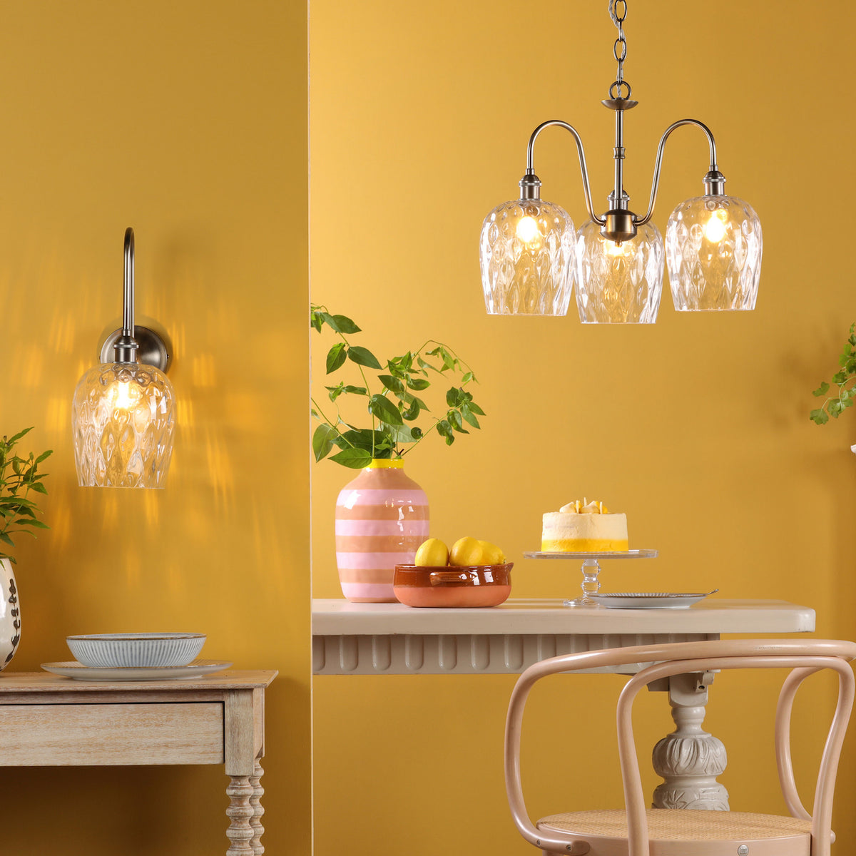A dining room with yellow walls, a wall sconce, and a chandelier. A table with lemons and a cake.