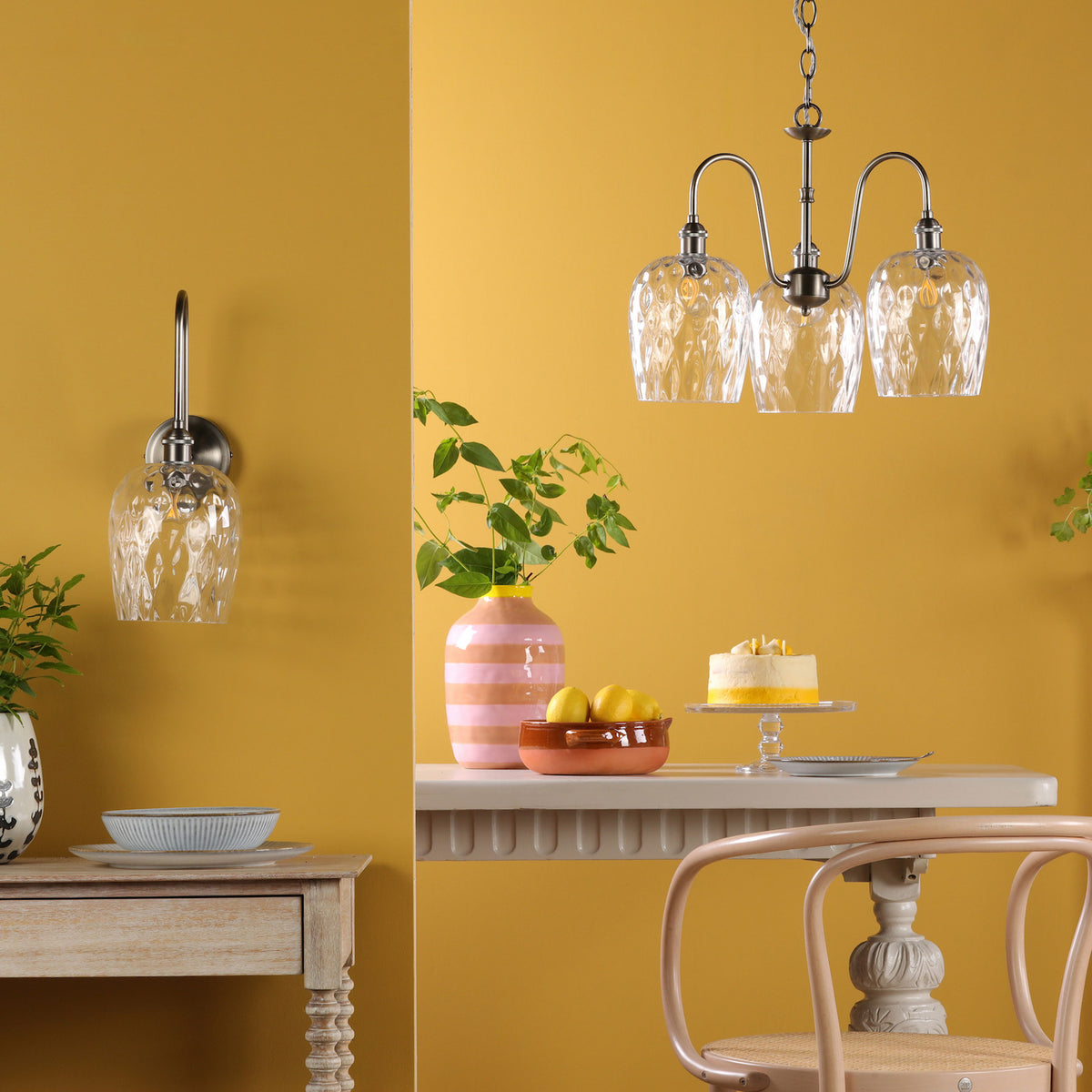 A room with yellow walls, a wall sconce, a chandelier, a table with a cake, lemons, and a chair.