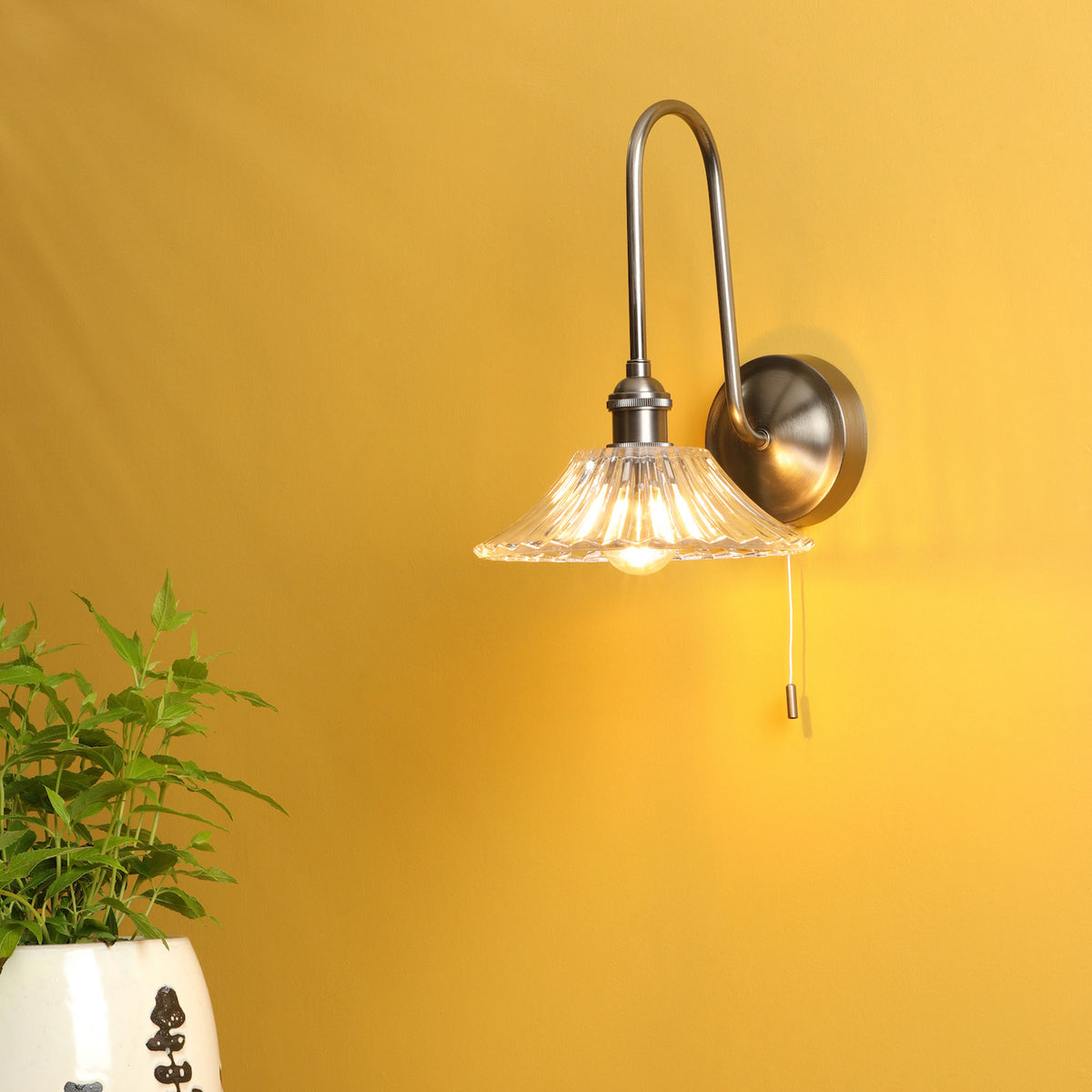 A brushed nickel wall sconce with a fluted glass shade illuminates a yellow wall, next to a potted plant.
