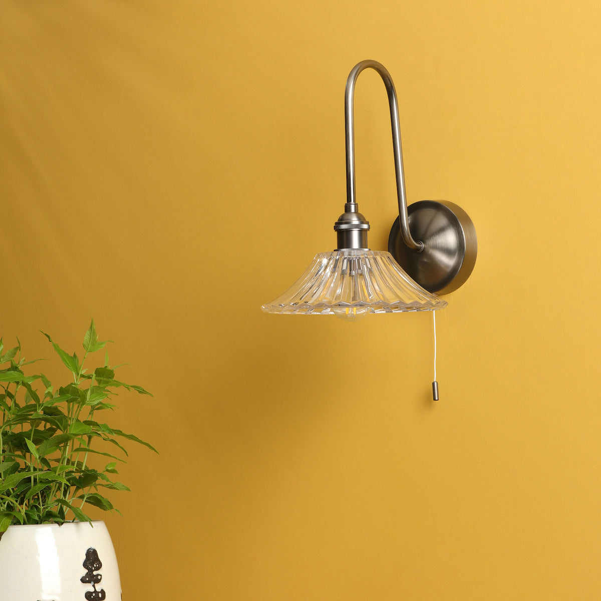 A brushed nickel wall sconce with a clear, fluted glass shade and a pull chain switch, mounted on a yellow wall.