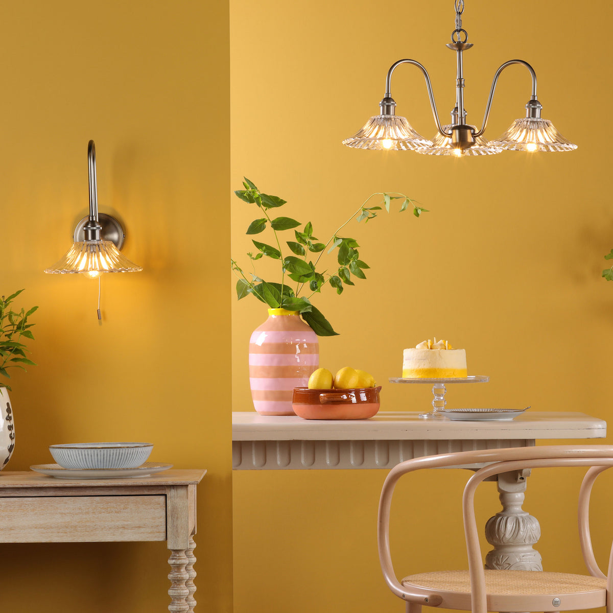 A dining room with yellow walls, a wall light, a chandelier, a table with a cake, and a chair.