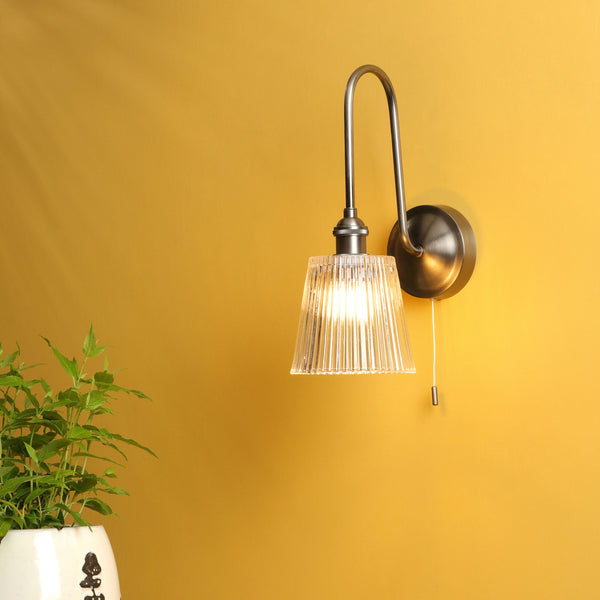 A wall-mounted light fixture with a ribbed glass shade and a pull chain, next to a potted plant.