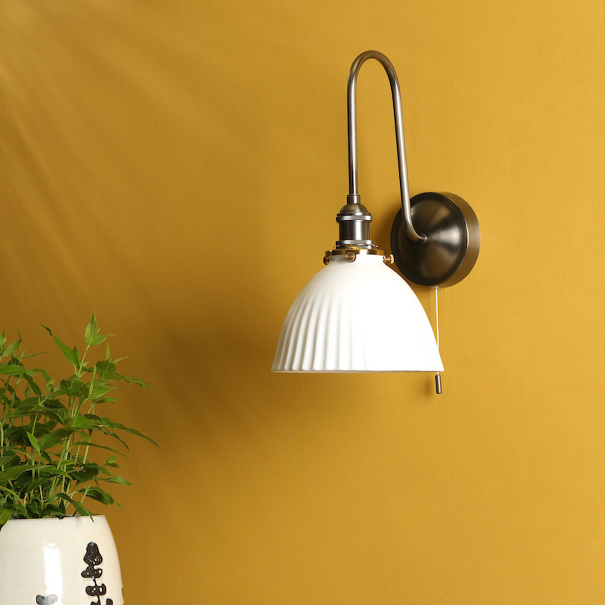 A white fluted wall sconce with a brushed metal arm and base, mounted on a mustard yellow wall.