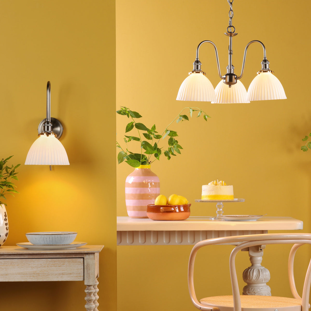 A wall light and a three-arm chandelier with pleated shades illuminate a yellow room with a table and chair.